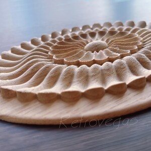 K&ouml;nnte beinhalten: Ein geschnitztes dekoratives Element aus Holz mit floralem Design. Das St&uuml;ck zeigt eine zentrale Bl&uuml;tenform, die von geschichteten, wellenartigen Mustern umgeben ist. Das Holz hat eine nat&uuml;rliche, hellbraune Farbe und ist auf einem runden Sockel montiert.
