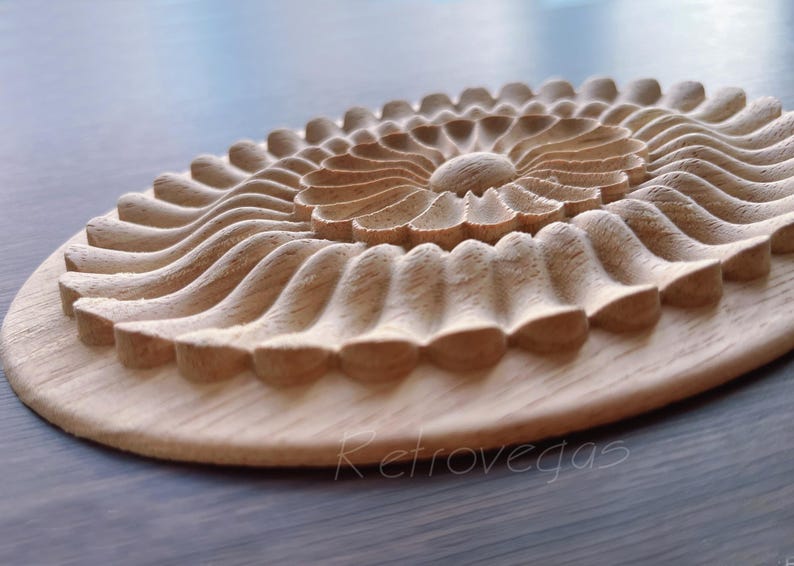 K&ouml;nnte beinhalten: Ein geschnitztes dekoratives Element aus Holz mit floralem Design. Das St&uuml;ck zeigt eine zentrale Bl&uuml;tenform, die von geschichteten, wellenartigen Mustern umgeben ist. Das Holz hat eine nat&uuml;rliche, hellbraune Farbe und ist auf einem runden Sockel montiert.