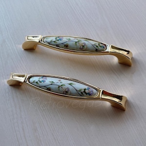 Może przedstawiać: Dwa złote uchwyty do szafek z wkładką z białej ceramiki z kwiatowym wzorem. Uchwyty mają owalny kształt i złote wykończenie.