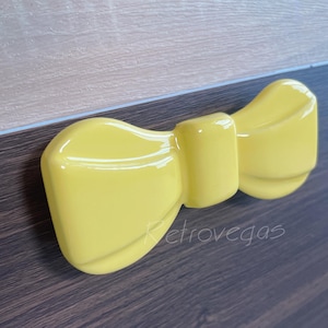 May include: A glossy, yellow bow-tie shaped decorative object. The bow has a smooth, reflective surface and is positioned on a dark wood-grain surface. The word "Retrovegas" is faintly visible on the object.