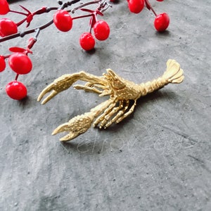 May include: A gold-colored crayfish brooch with detailed claws and a textured body. The brooch is lying on a gray textured surface.