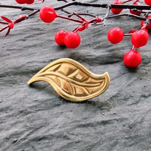 May include: A gold leaf-shaped brooch with a detailed design. The brooch is on a grey textured surface.