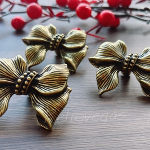 Op de afbeelding: Drie antiek goudkleurige, strikvormige decoratieve hardware-onderdelen. De strikken hebben gedetailleerde, getextureerde oppervlakken en zijn gerangschikt op een houten oppervlak. Rode bessen en takken staan op de achtergrond.