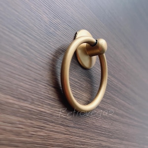 Könnte beinhalten: Goldener Ring-Griff für eine Schranktür mit runder Basis. Der Griff ist auf einer dunklen Holzoberfläche montiert.