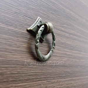 Op de afbeelding: Antieke bronzen ladeknop met een ruitvormige achterplaat en een decoratieve ringgreep. De handgreep heeft een gevlochten ontwerp. De hardware is gemonteerd op een donkerbruin houten oppervlak.