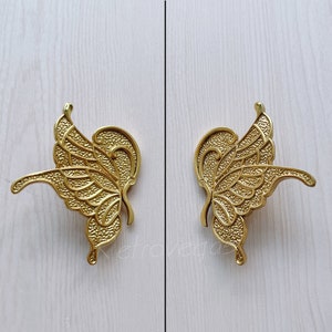 May include: A pair of gold butterfly-shaped cabinet door pulls. The pulls have a detailed, textured design.