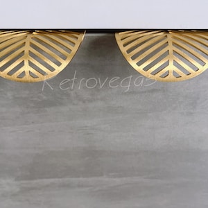 May include: Two gold-coloured leaf-shaped drawer pulls are mounted on a grey surface. The pulls have a cut-out design with a leaf vein pattern. The word "Retrovegas" is faintly visible in the background.