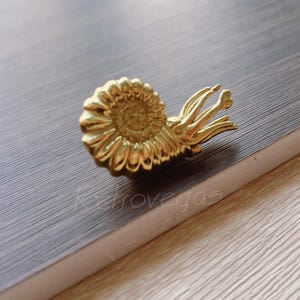 May include: A gold-coloured, detailed ammonite-shaped decorative object. The object is made of metal and has a spiral design with intricate detailing. The object is resting on a dark grey surface. The word "Katrovegas" is visible in the image.