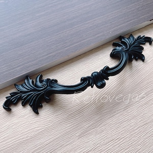 May include: A black, ornate cabinet pull with a curved handle and decorative leaf-like details. The hardware is made of metal and is designed for furniture. The pull is shown on a light wood surface.