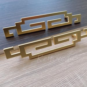 Op de afbeelding: Twee gouden metalen ladegrepen met een geometrisch ontwerp. De grepen zijn gevormd als een gestileerde wolk met een herhalend patroon.