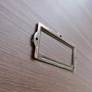 May include: An antique brass label holder, rectangular in shape, with two small mounting holes. It is set against a dark wood grain background. The metal has a vintage, aged appearance.