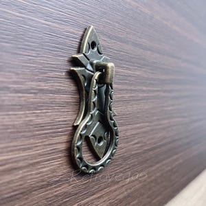 May include: Antique-style bronze drawer pull with a decorative, ornate design. The pull has a loop handle and a pointed top.
