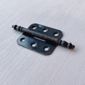 May include: A black metal hinge with decorative accents. The hinge has a square base and a curved arm with a decorative knob on each end.