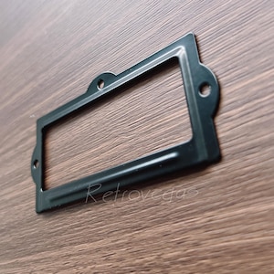 以下が含まれることがあります： 丸みを帯びたエッジと長方形の開口部を持つ黒い金属製のラベルホルダー。ホルダーは表面に取り付けるように設計されています。