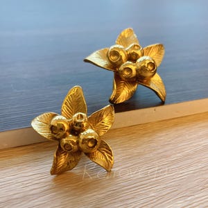 May include: Two gold-colored flower-shaped earrings. Each earring features five petals and a cluster of small, round accents in the center. The earrings are displayed on a wooden surface with a blue background.
