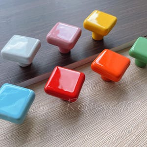 Może przedstawiać: Zestaw kolorowych kwadratowych gałek ceramicznych do szafek. Gałki są w odcieniach niebieskiego, zielonego, ż&oacute;łtego, pomarańczowego, czerwonego, r&oacute;żowego i białego.