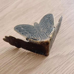 Puede incluir: Un soporte de esquina de metal dorado con un diseño de mariposa. El soporte es ornamentado y tiene un diseño detallado.