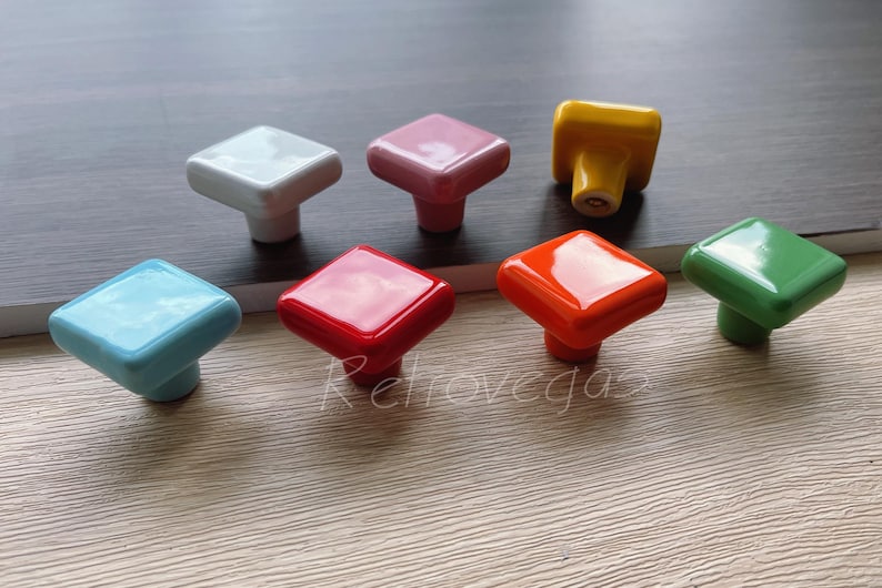 Może przedstawiać: Zestaw ośmiu kwadratowych ceramicznych gałek do szafek w r&oacute;żnych kolorach, w tym biały, r&oacute;żowy, ż&oacute;łty, pomarańczowy, czerwony, niebieski, zielony i jasnoniebieski.