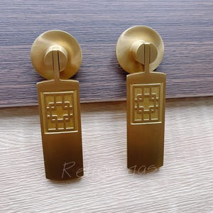 Peut inclure: Deux poignées de meuble en laiton avec un motif géométrique. Chaque poignée a une plaque arrière circulaire, un corps rectangulaire avec un motif carré et une barre de connexion. Les poignées sont de couleur or chaud.