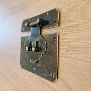 Puede incluir: Pestillo de latón antiguo con un diseño decorativo. El pestillo tiene dos partes, una parte superior y una parte inferior, que están conectadas por una varilla de metal. El pestillo está en un acabado desgastado.