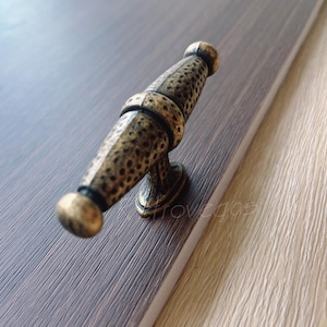 May include: Antique bronze cabinet pull with a textured, cylindrical design. The hardware is mounted on a dark wood surface, showcasing its decorative details and vintage aesthetic. The pull features a detailed, hammered texture.