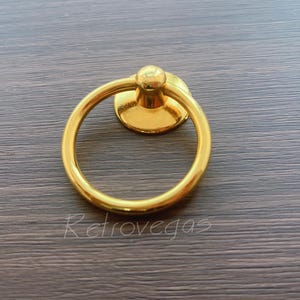 Könnte beinhalten: Ein goldfarbener Ringgriff mit rundem Griff und kleinem, rundem Sockel. Der Griff ist auf einer kreisförmigen Platte montiert und hat eine kleine, dekorative Kugel oben. Das Wort "Retrovegas" ist auf der Oberfläche aufgedruckt.