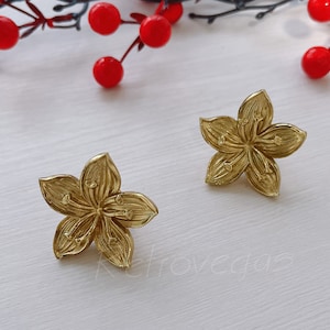 May include: Two gold-colored flower-shaped earrings. Each earring has five petals and detailed stamens. The earrings are on a white surface with red berries and a dark green stem in the background. The word "retrovegas" is in the bottom right corner.