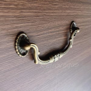 May include: Antique-style brass drawer pull with a decorative design. The pull has a curved shape and a rounded end.