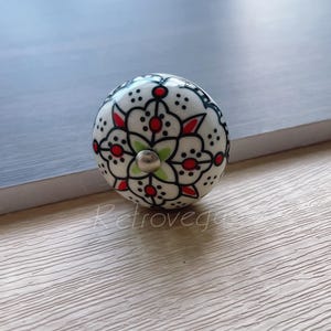 Peut inclure: Bouton en céramique blanche avec un motif floral noir et rouge. Le bouton a une vis en métal argenté au centre.