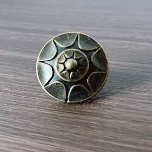 May include: A round, antique brass-colored cabinet knob with a starburst design. The knob features a central, raised circle surrounded by stylized petal shapes. The hardware is set against a dark wood grain surface.