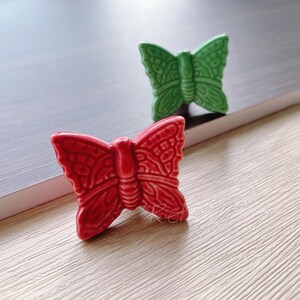 Könnte beinhalten: Zwei Keramik-Schmetterlingsfiguren. Eine ist leuchtend rot, die andere ist ein sattes Grün. Beide haben detaillierte Flügelmuster und sind auf einer hellen Oberfläche platziert, was auf ein dekoratives oder künstlerisches Thema hindeutet.