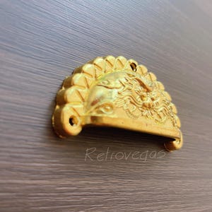 May include: Gold-toned metal drawer pull with a decorative design featuring a stylized dragon head. The pull is shaped like a half-circle.