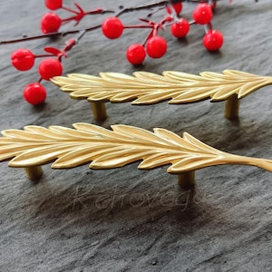 Könnte beinhalten: Zwei goldene, blattförmige Schrankgriffe mit einem detaillierten Design. Die Griffe sind aus Metall und haben eine polierte Oberfläche.