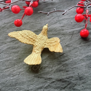 May include: A gold-colored metal dove figurine with its wings spread out. The dove is sitting on a gray textured surface.
