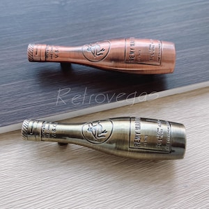 May include: Two antique-style bottle stoppers shaped like champagne bottles. The top bottle stopper is copper-coloured with the text "REMY MARTIN VSOP" and the bottom bottle stopper is a greenish-gold colour with the text "REMY MARTIN VSOP Cognac Fine Champagne".