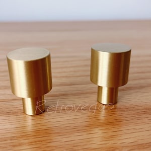 May include: Two gold cabinet knobs with a cylindrical design. The knobs have a rounded top and a smaller, cylindrical base. They are set against a light brown wooden surface.