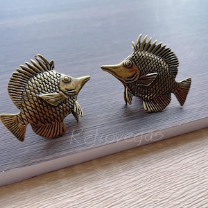 May include: Two gold-coloured metal fish figurines with detailed scales and fins. The fish are facing each other and have a slightly raised, three-dimensional appearance.