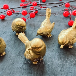 May include: Five gold-coloured bird figurines, each with a different pose, are arranged on a grey textured surface. The birds are made of a smooth, shiny material and have a realistic appearance.