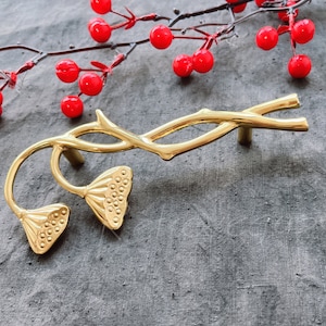 May include: A gold-colored cabinet pull with a floral design. The pull features a branch-like handle and two lotus flower-shaped ends. Red berries on a branch are in the background. The hardware is likely for a cabinet or drawer.
