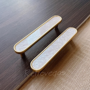 May include: Two white and gold cabinet pulls with a marbled design. The pulls are oval shaped and have a brass finish.