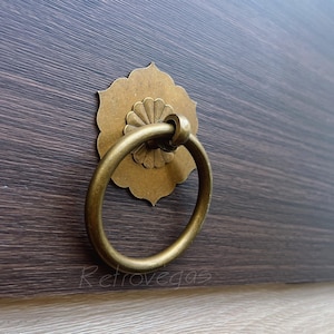 May include: A brass drawer pull with a ring handle, set against a dark brown wooden surface. The pull features a floral backplate. The hardware has an antique finish. The wood grain is visible.