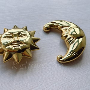 May include: Gold-toned metal sun and moon charms with detailed facial features. The sun has a round face with a smile and the moon has a crescent shape with a face.
