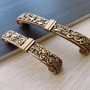 Può includere: Due tiretti per mobili in tonalità oro, decorati con un motivo floreale. I tiretti sono curvi e presentano un design leggermente in rilievo.