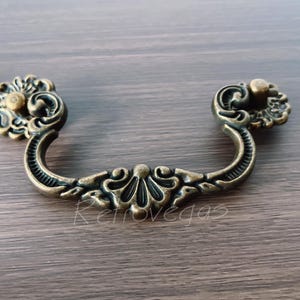 Könnte beinhalten: Antiker Messing-Schubladengriff mit aufwendigen floralen Details. Der Griff hat ein geschwungenes Design mit dekorativen Endstücken und einem zentralen floralen Akzent. Ein Hardware-Teil im Vintage-Stil.