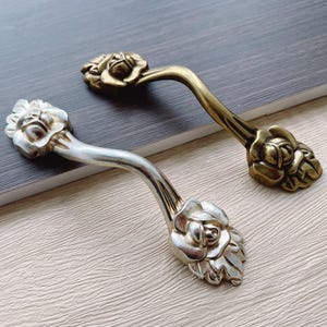 Może przedstawiać: Dwa ozdobne uchwyty do szafek z motywem róży. Jeden uchwyt jest srebrny, a drugi złoty.