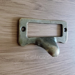May include: A vintage-style brass label holder with a pull handle. The rectangular frame has two screw holes and a curved handle. The metal has an aged, tarnished appearance, set against a wood grain background.
