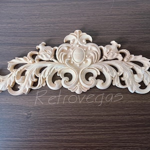 Puede incluir: Moldura arquitectónica de madera crema ornamentada con un diseño simétrico. La moldura presenta hojas en espiral, elementos florales y una forma ovalada central. La palabra "Retrovegas" está escrita en la parte inferior.