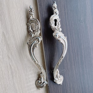 May include: Two ornate silver door handles with a keyhole in the centre. The handles are intricately designed with swirling patterns and a decorative finish.