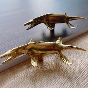 May include: Two gold-toned dinosaur-shaped cabinet knobs. The knobs are detailed with scales and fins.
