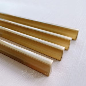 以下が含まれることがあります： 斜めに配置された4つの金色のキャビネットプル。各プルは、湾曲した凹型ハンドルを備えた長方形の形状をしています。プルは光沢のある金属素材でできており、明るい色の木材の表面に配置されています。